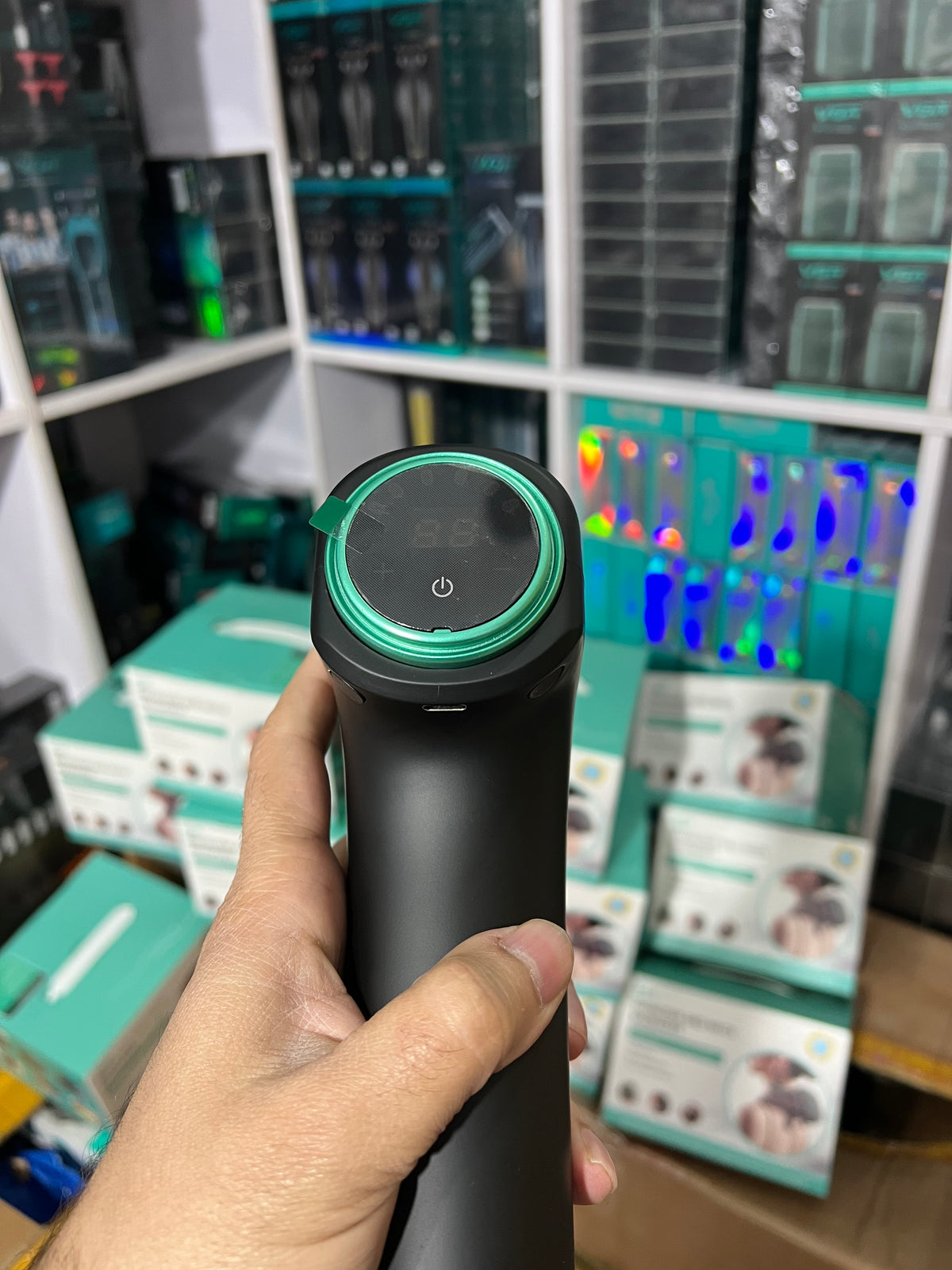 Hand holding a black and green cylindrical device with blurred electronic components in the background