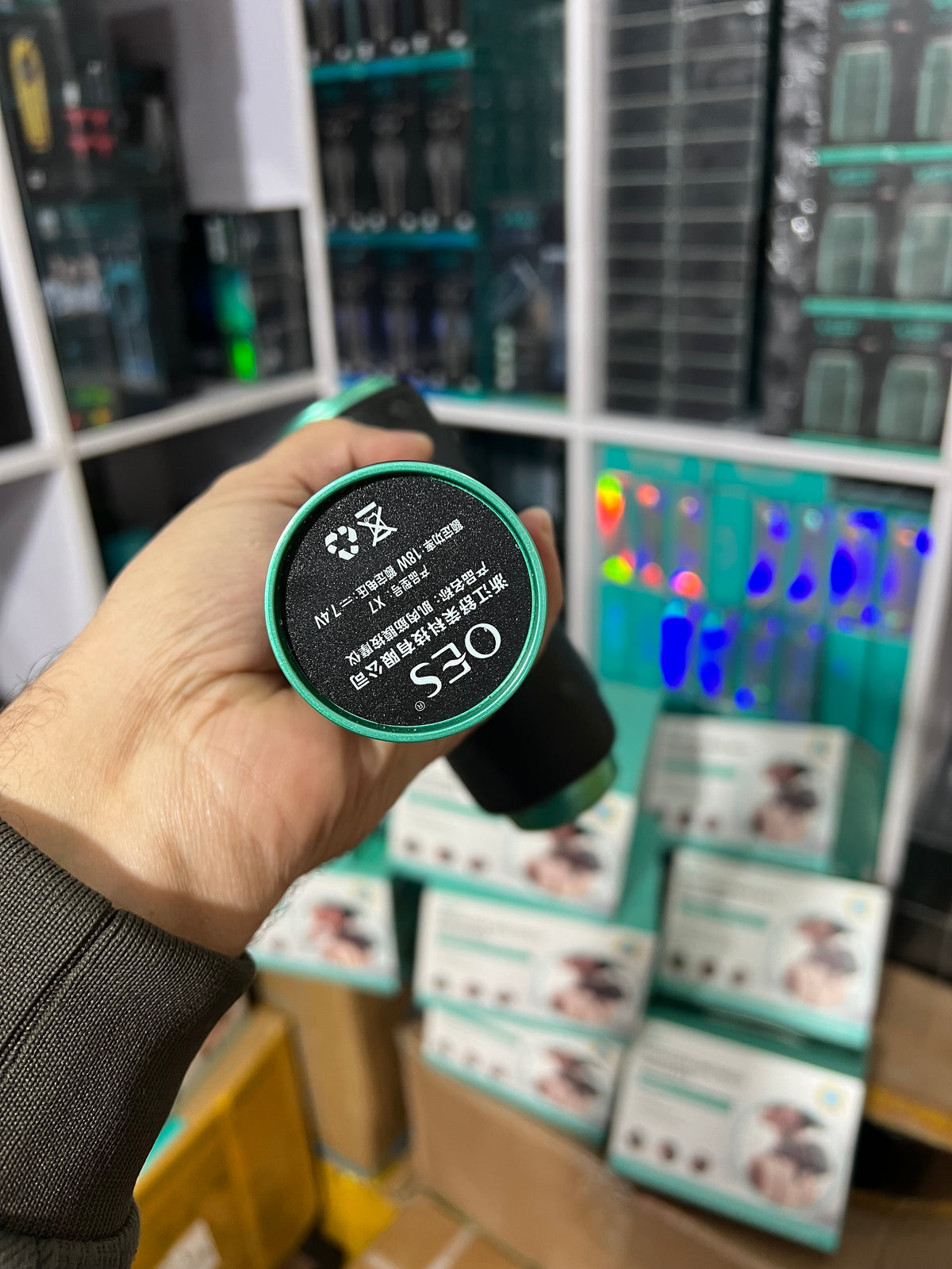 Hand holding a device with 'OES' branding in front of electronic equipment and displays.