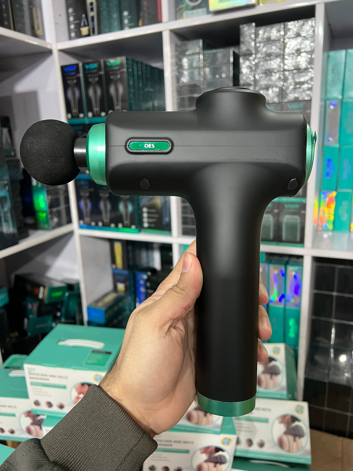 Hand holding a black and green massage gun in front of a store shelf.